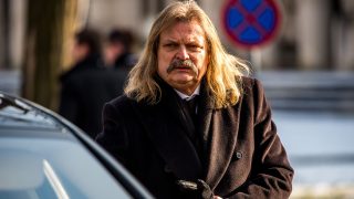 Man with long blond hair and a mustache, wearing a dark overcoat, standing beside a car on a city street.