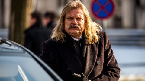 Man with long blond hair and a mustache, wearing a dark overcoat, standing beside a car on a city street.