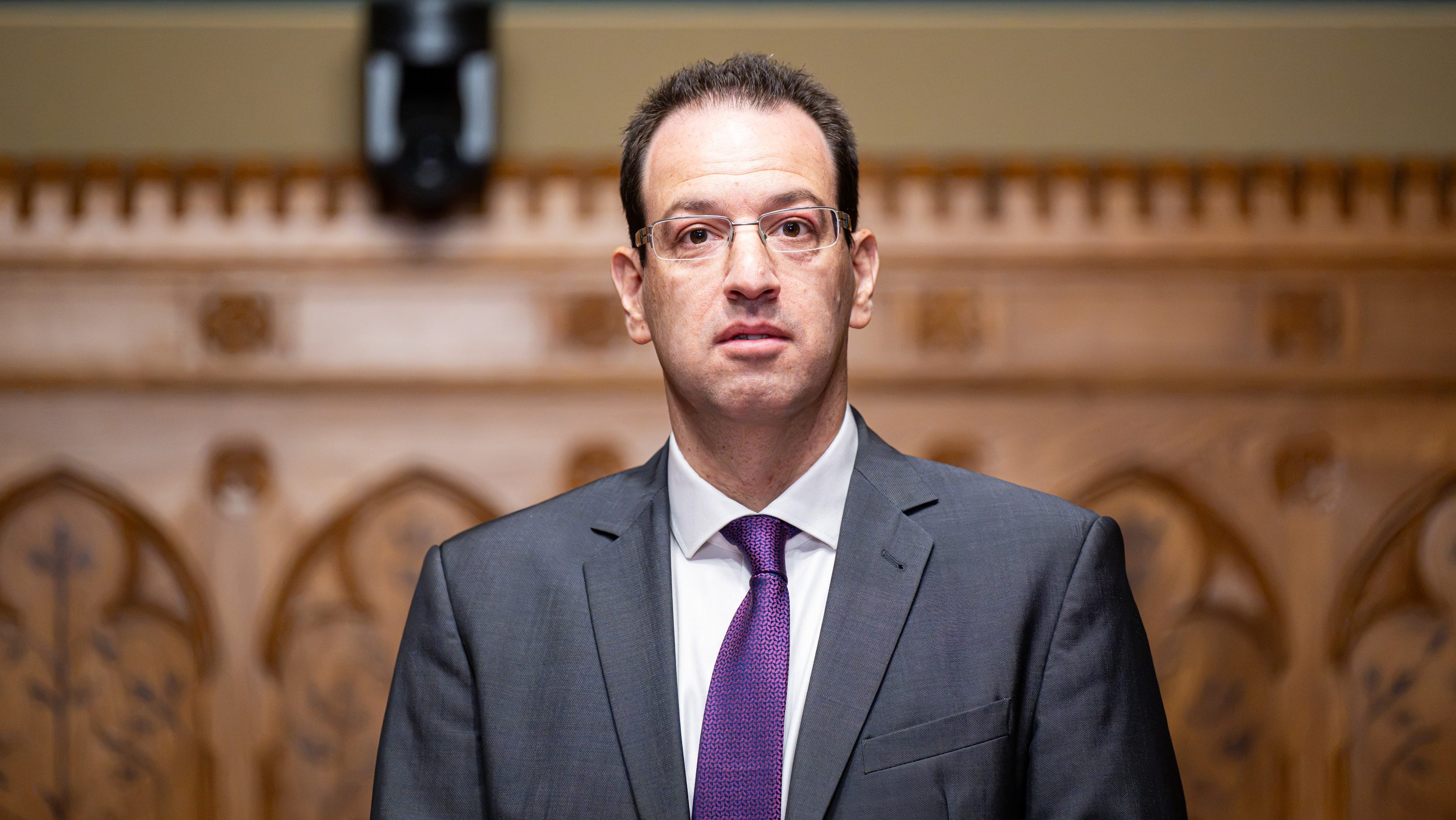 Man in a dark gray suit and purple tie posing in a wood-paneled auditorium, looking at the camera, with blurred rows behind him.