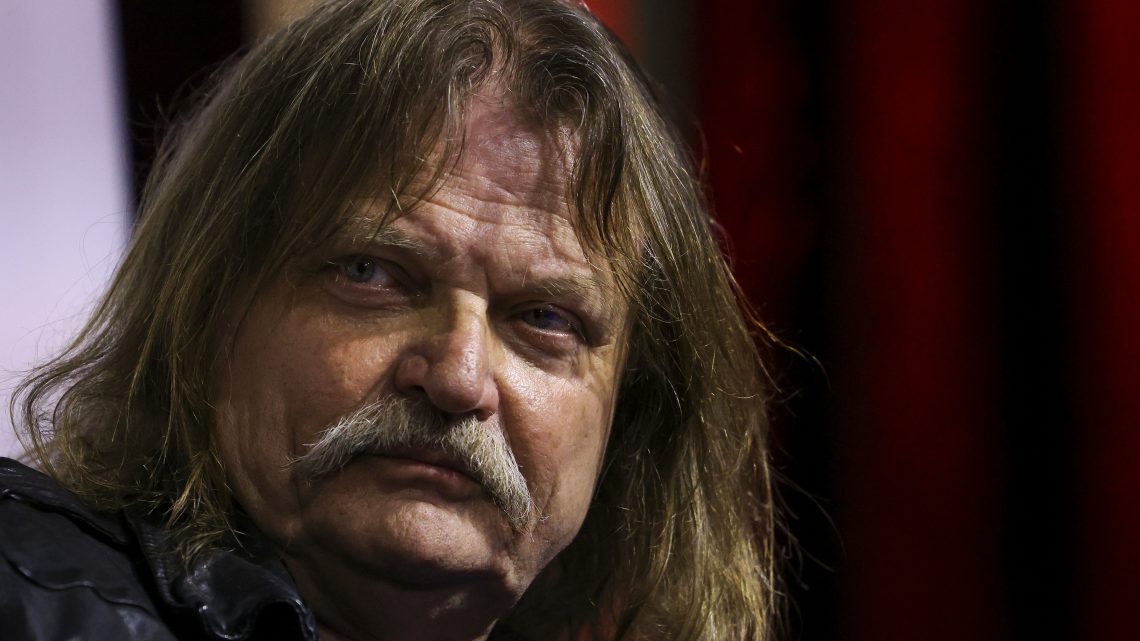 Older man with long, gray-blond hair and a thick mustache looks toward the camera, with a neutral expression.