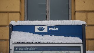 Blue MÁV railway sign with the Hungarian rail network text, topped with snow, against a yellow building backdrop.