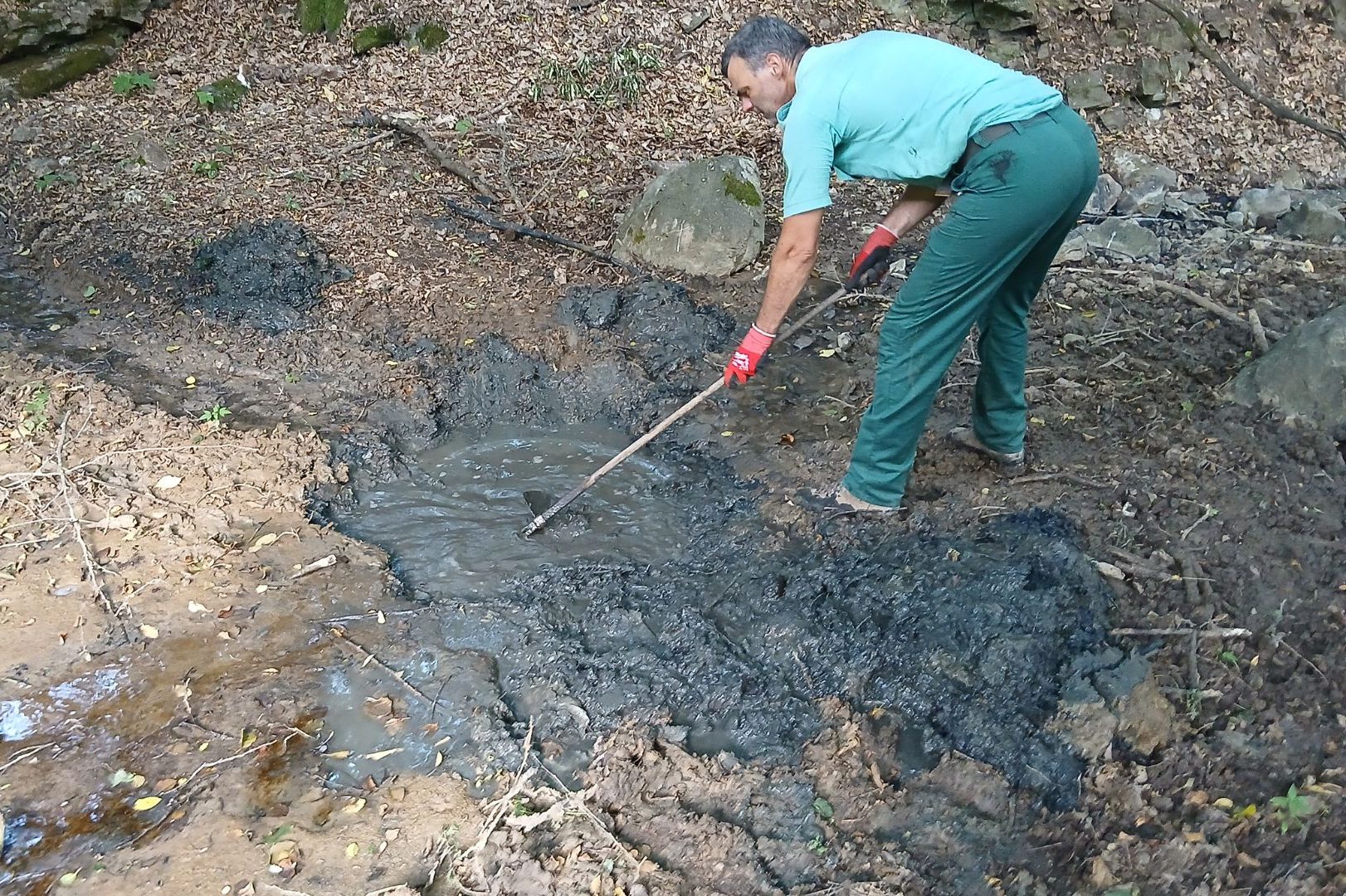 Man in a teal shirt and green pants shovels muddy water from a dark, muddy pit in a forested area.