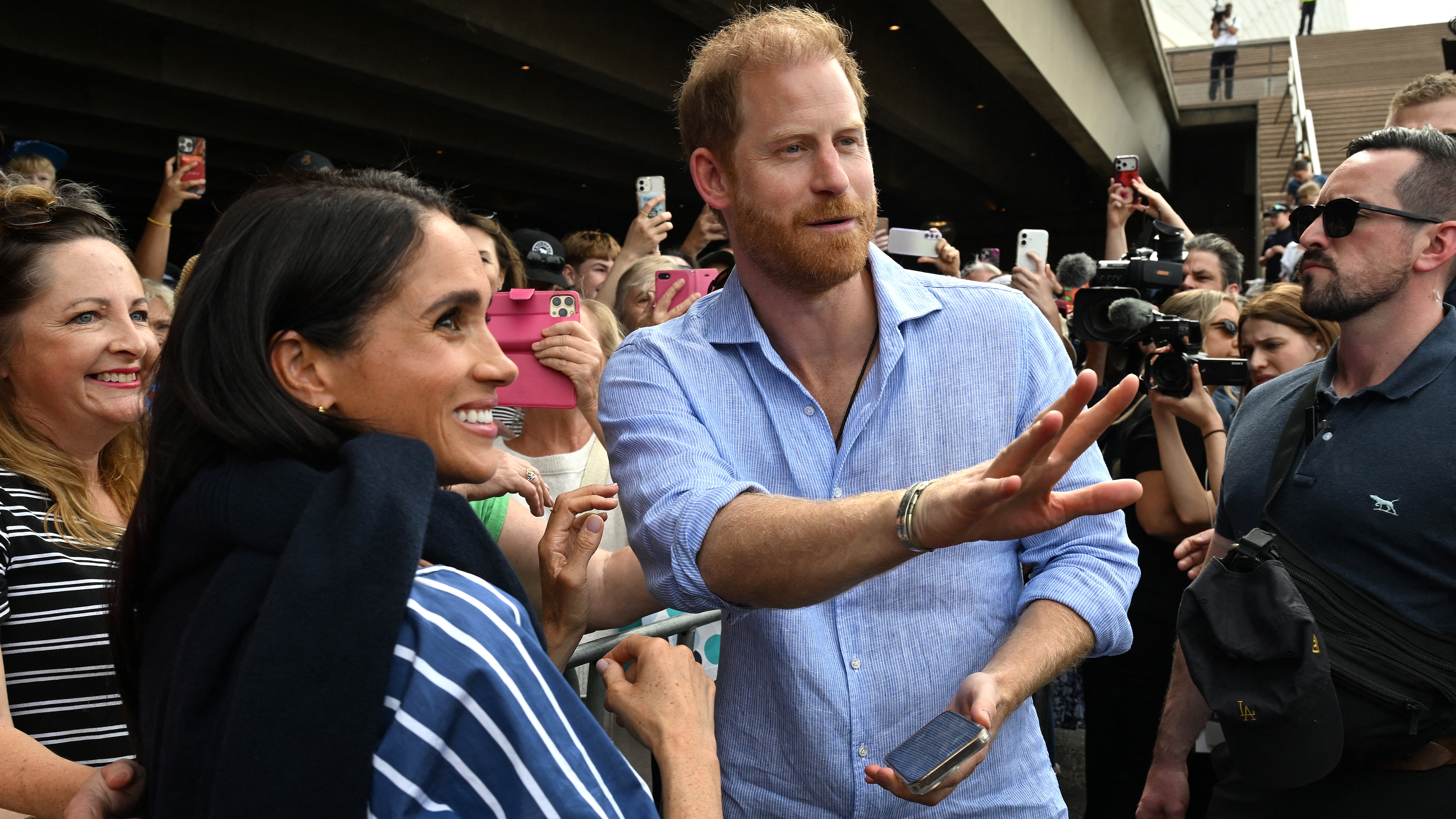 Prince Harry in a light blue shirt greeting fans as a crowd of people hold up phones and cameras around him.