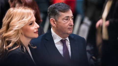 Man in a dark suit and purple tie sitting beside a blonde woman at a formal event, with a crowd in the background, both looking ahead.