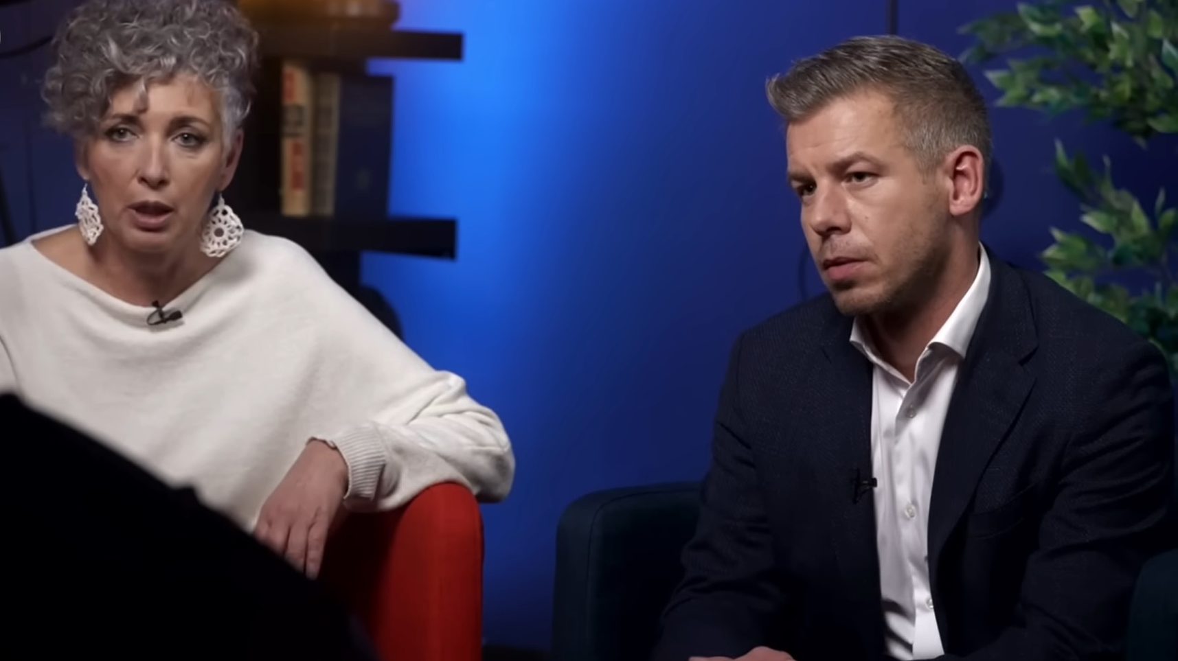 Two people sit in a studio setup: a woman with short curly gray hair and white sweater speaking, wearing large white earrings; a man in a dark blazer listens on her right.
