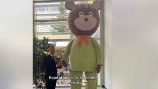 Man in a suit hands a trophy to a giant bear mascot in a lime-green outfit indoors by large windows.