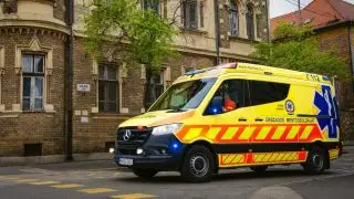 Yellow ambulance with blue lights driving past a brick historic building on a city street in Hungary.