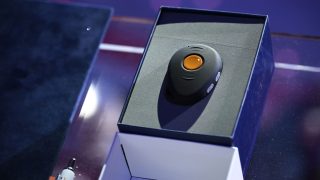 Close-up of a black circular gadget with an orange center button in its open box on a dark surface.