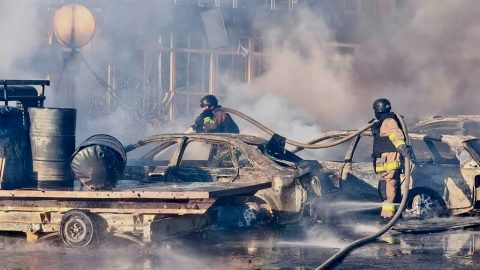 Firefighters using hoses to extinguish a fire among burnt cars in an industrial lot at a smoky scene.