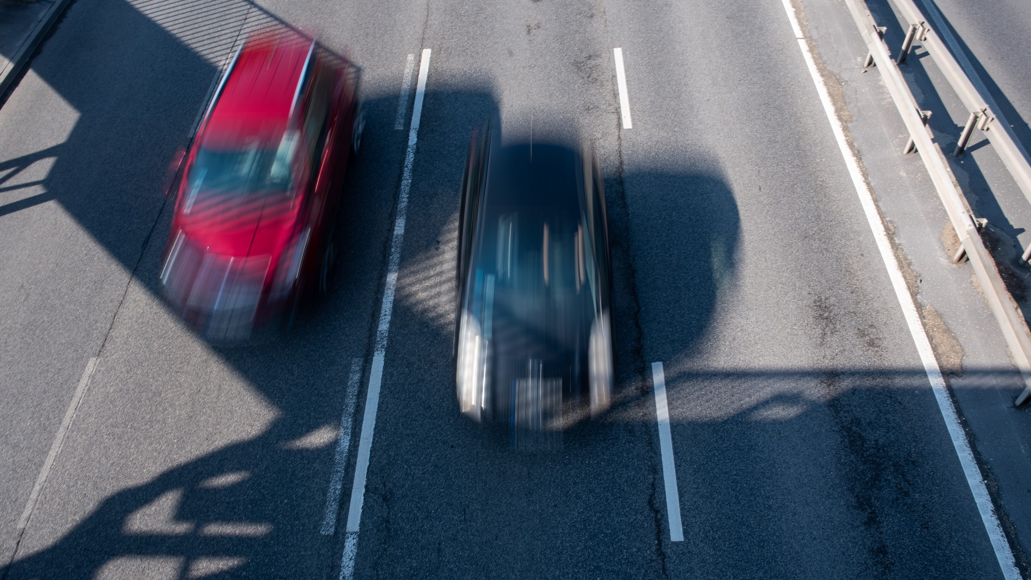 Aerial view of a highway with two speeding cars blurred by motion, one red on the left and a dark vehicle on the right, between white lane lines and a guardrail on the right side.