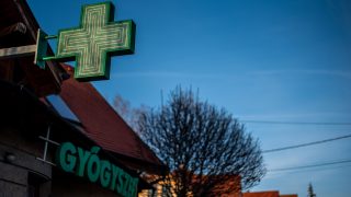 Green illuminated cross-shaped pharmacy sign mounted on the side of a building against a clear blue sky.