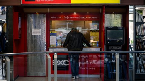 Man wearing a leather jacket stands at a red currency exchange booth, facing the window to exchange money, with multilingual 'PÉNZVÁLTÁS | CHANGE | GELDWECHSEL' sign above.