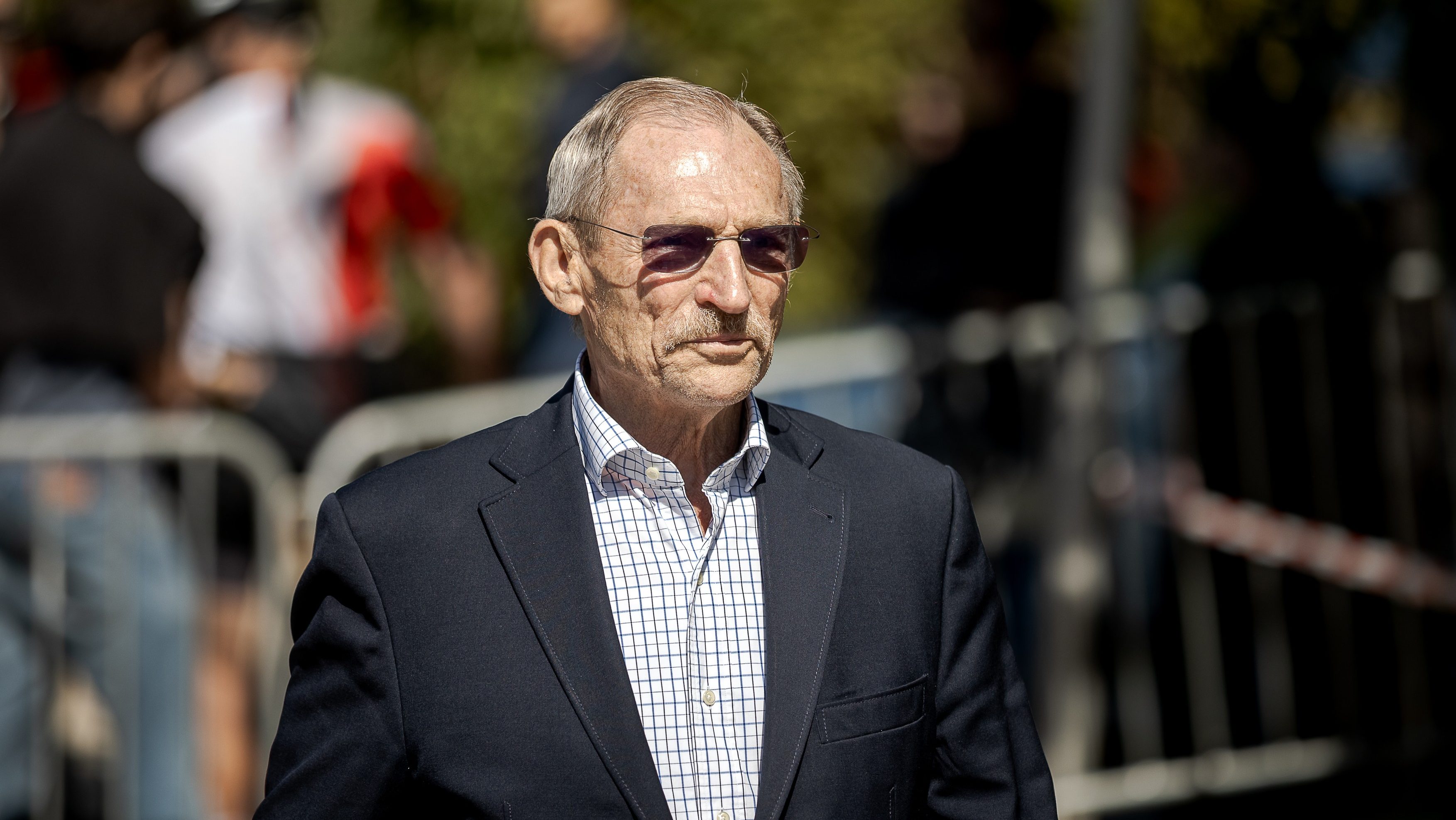 Older man in a navy blazer and checkered shirt wearing sunglasses at an outdoor event, with a blurred crowd behind him.