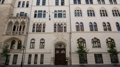 Facade of a pale stone historic building with tall arched windows and decorative Gothic detailing, central wooden doors, and a person in a hi-vis vest near the entrance