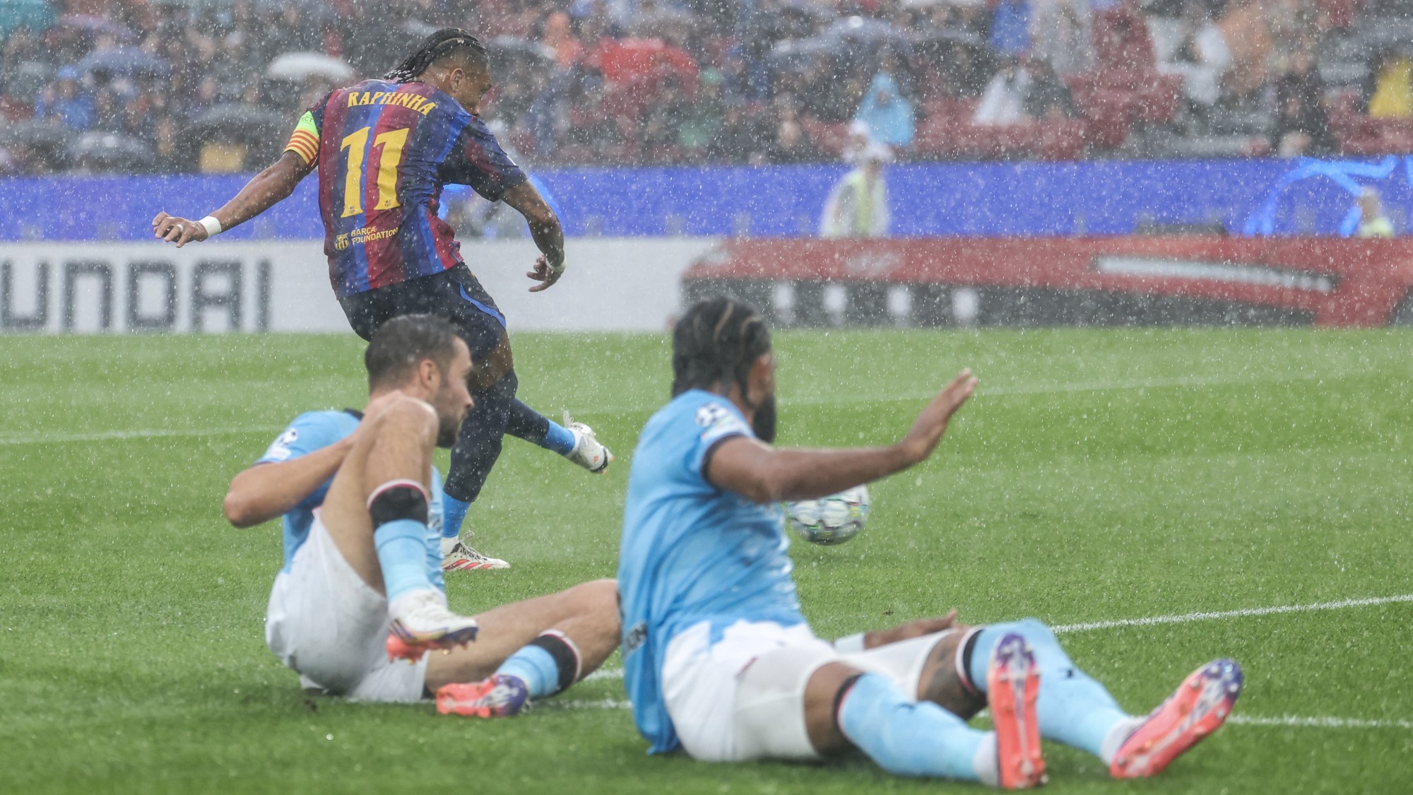 Soccer players slide-tackling in heavy rain on a green pitch; a defender in a maroon-and-blue striped jersey with the number 11 stands up toward the ball.