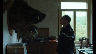Man in uniform stands in a dim room, looking at a large mounted bear head on the wall.