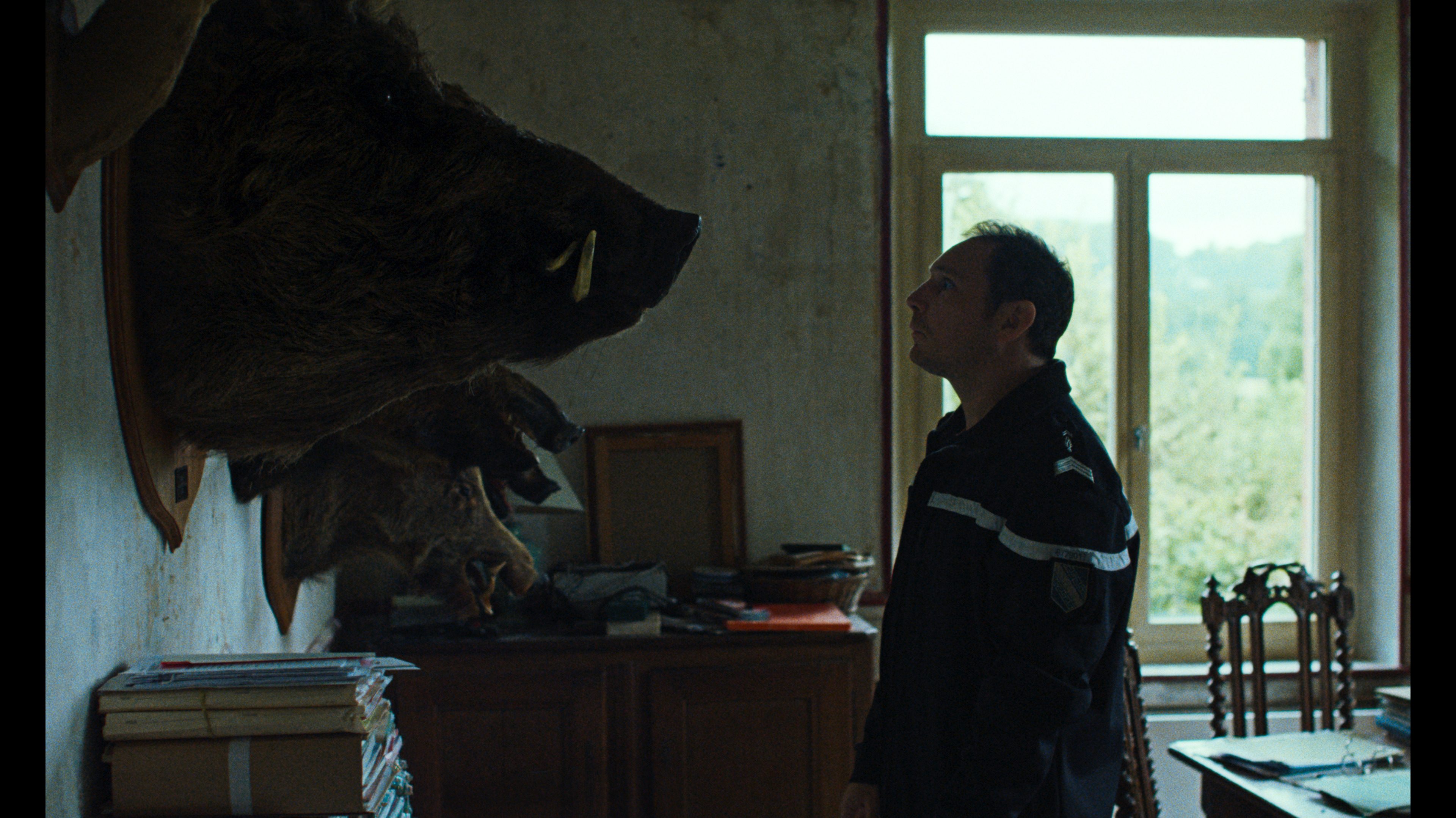 Man in uniform stands in a dim room, looking at a large mounted bear head on the wall.