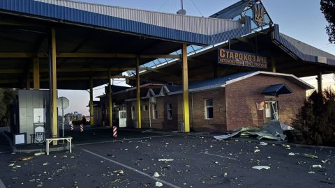 Border checkpoint under a large blue-and-metal canopy with yellow support columns; debris and torn panels are scattered across the asphalt.