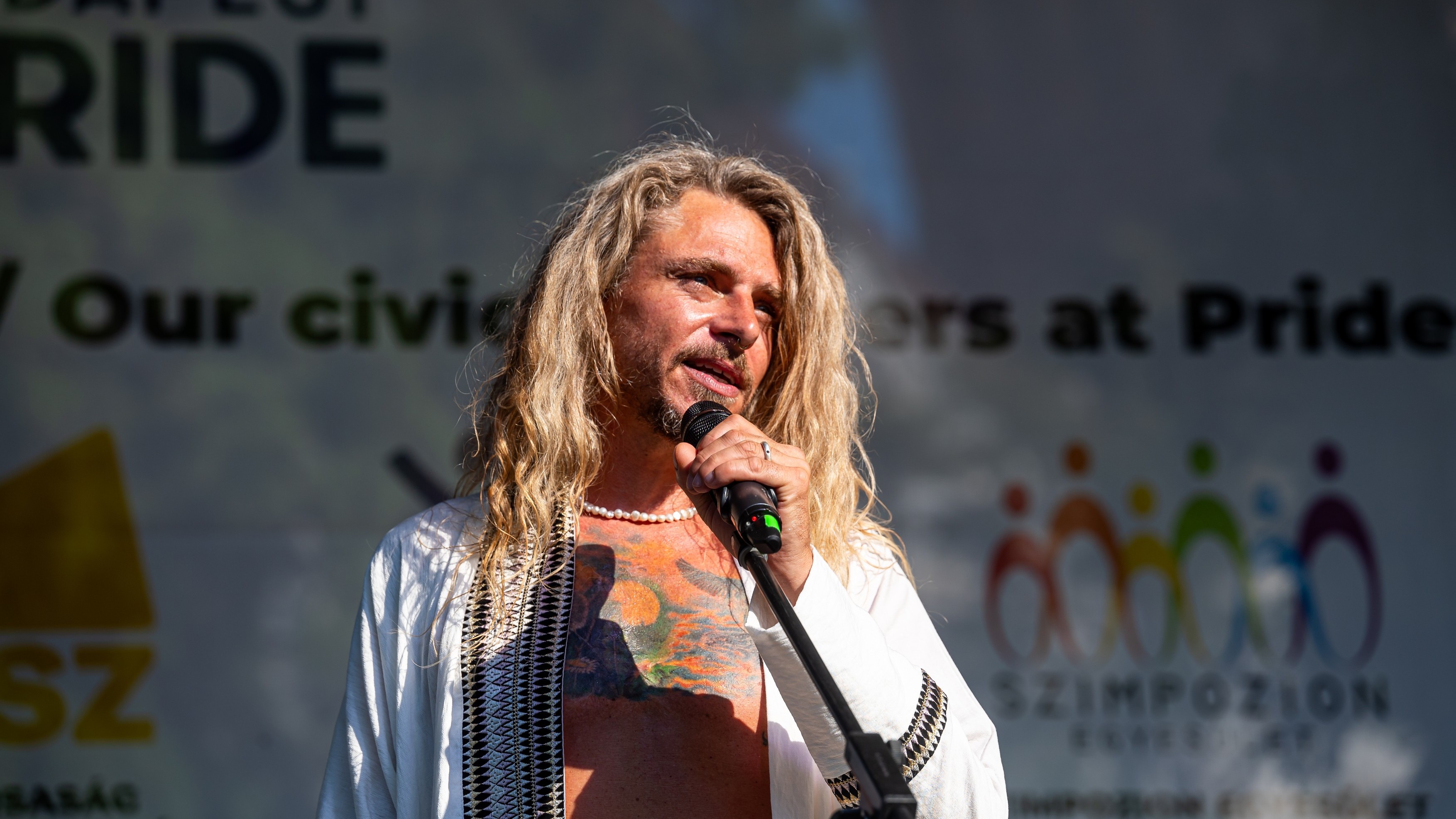 Male singer with long blond hair performing on stage, holding a microphone close to his mouth.