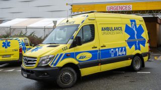 Yellow ambulance van parked outside a hospital entrance with URGENCES sign in the background; emergency medical vehicle ready for transport.
