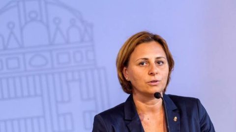 Woman in a navy blazer speaks into a microphone at a press conference, with a pale blue backdrop featuring a faint building outline.