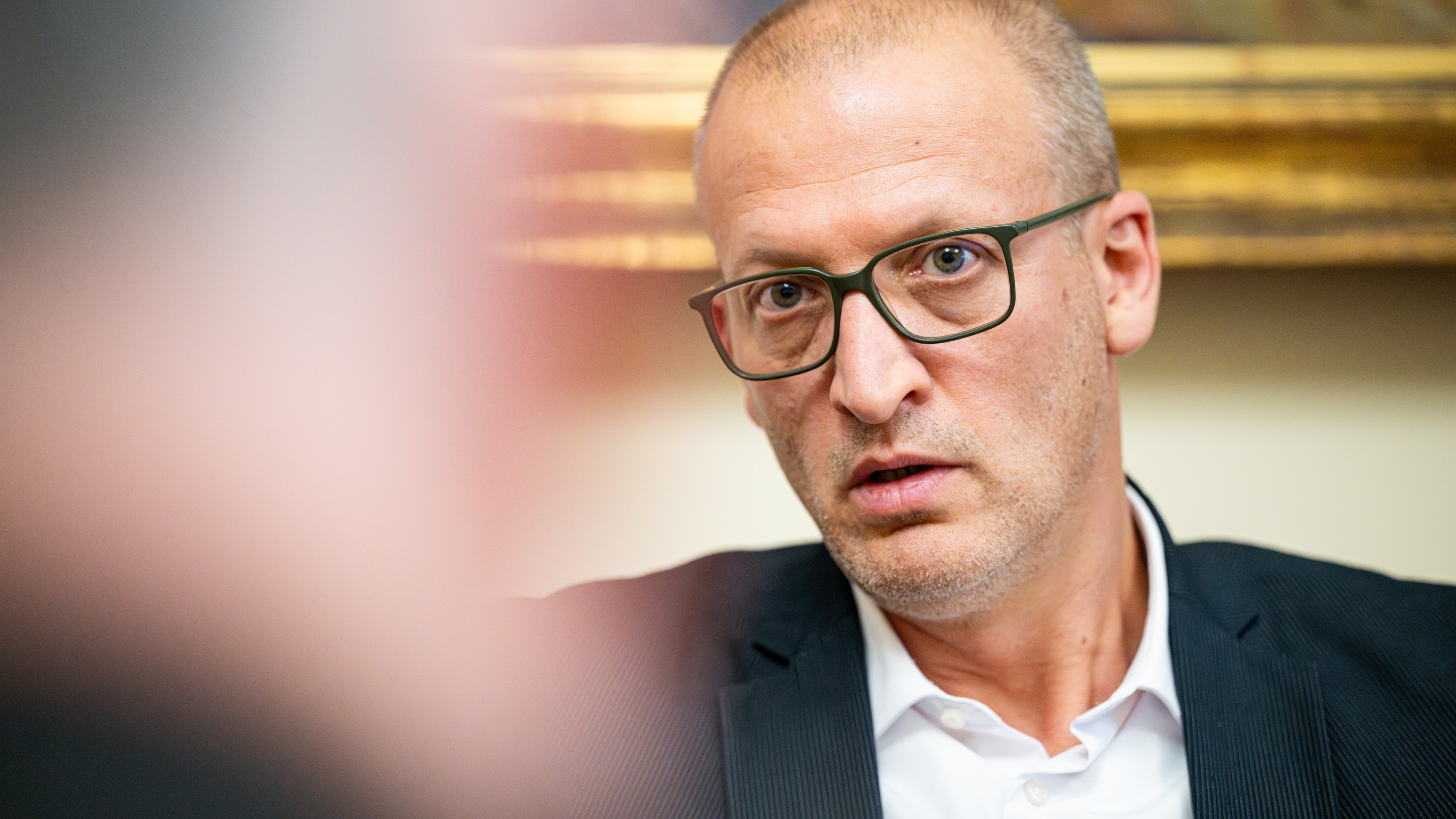 Close-up portrait of a man wearing dark-framed glasses in a suit, looking toward the camera.