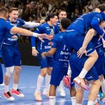 Blue-clad handball players celebrate together in a huddle on the court, as a cheering crowd looks on in the background.