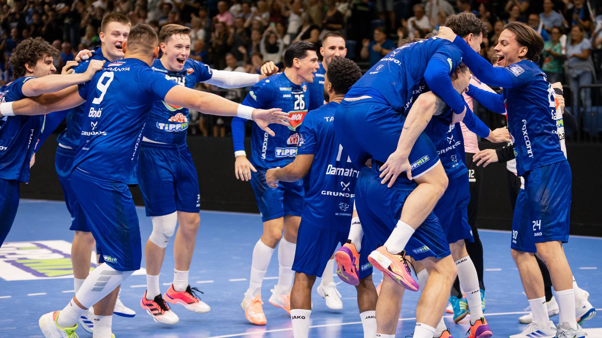 Blue-clad handball players celebrate together in a huddle on the court, as a cheering crowd looks on in the background.
