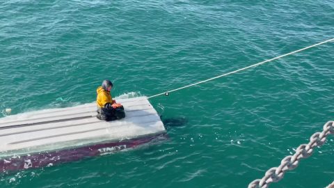 Person in a yellow rain jacket sits cross‑legged on a small floating platform, tethered by a rope to a boat in turquoise water.
