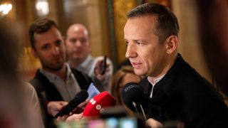 Man in a dark jacket speaks to reporters at a press event with multiple microphones in front of him