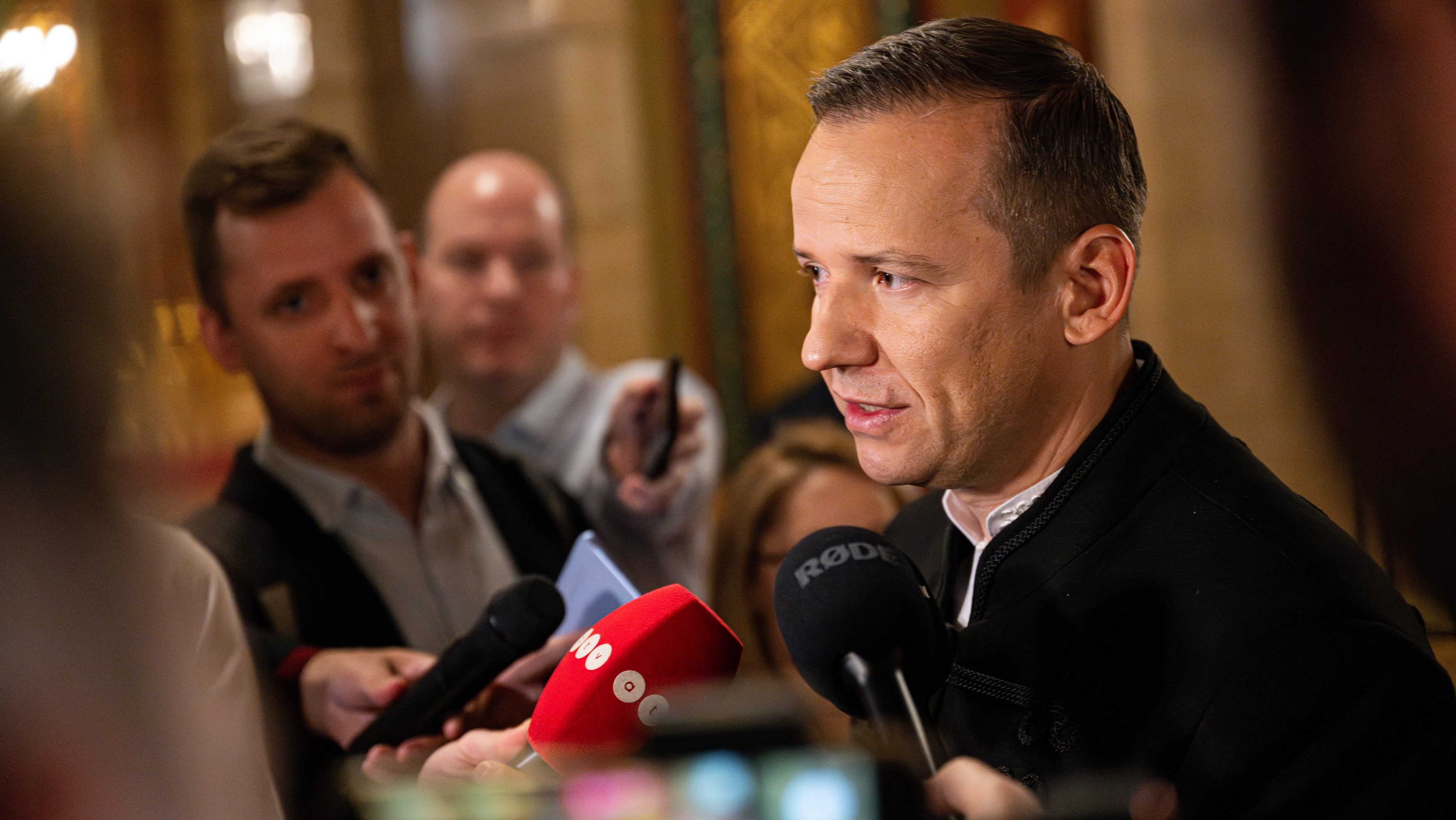 Man in a dark jacket speaks to reporters at a press event with multiple microphones in front of him