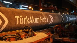Industrial pipe labeled 'TürkAkım Hattı' in a machinery-filled factory setting, orange-hued lighting