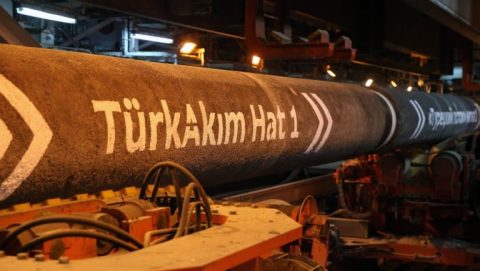 Industrial pipe labeled 'TürkAkım Hattı' in a machinery-filled factory setting, orange-hued lighting