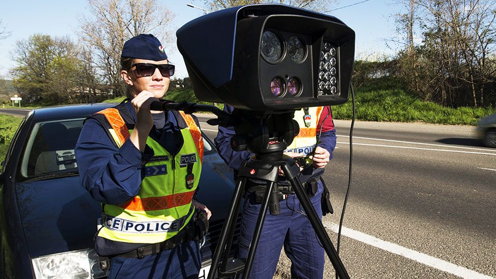 Two police officers set up a speed camera on a tripod by the roadside, wearing reflective vests and dark uniforms.