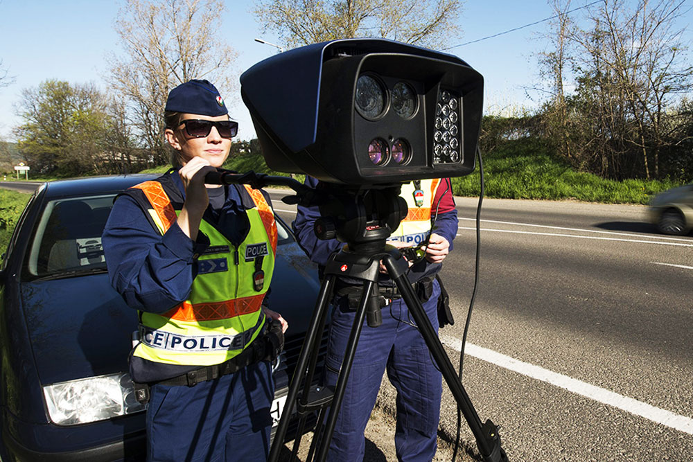 Two police officers set up a speed camera on a tripod by the roadside, wearing reflective vests and dark uniforms.