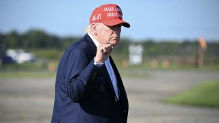 Man in a dark suit outdoors, wearing a red MAGA cap and making a fist gesture.
