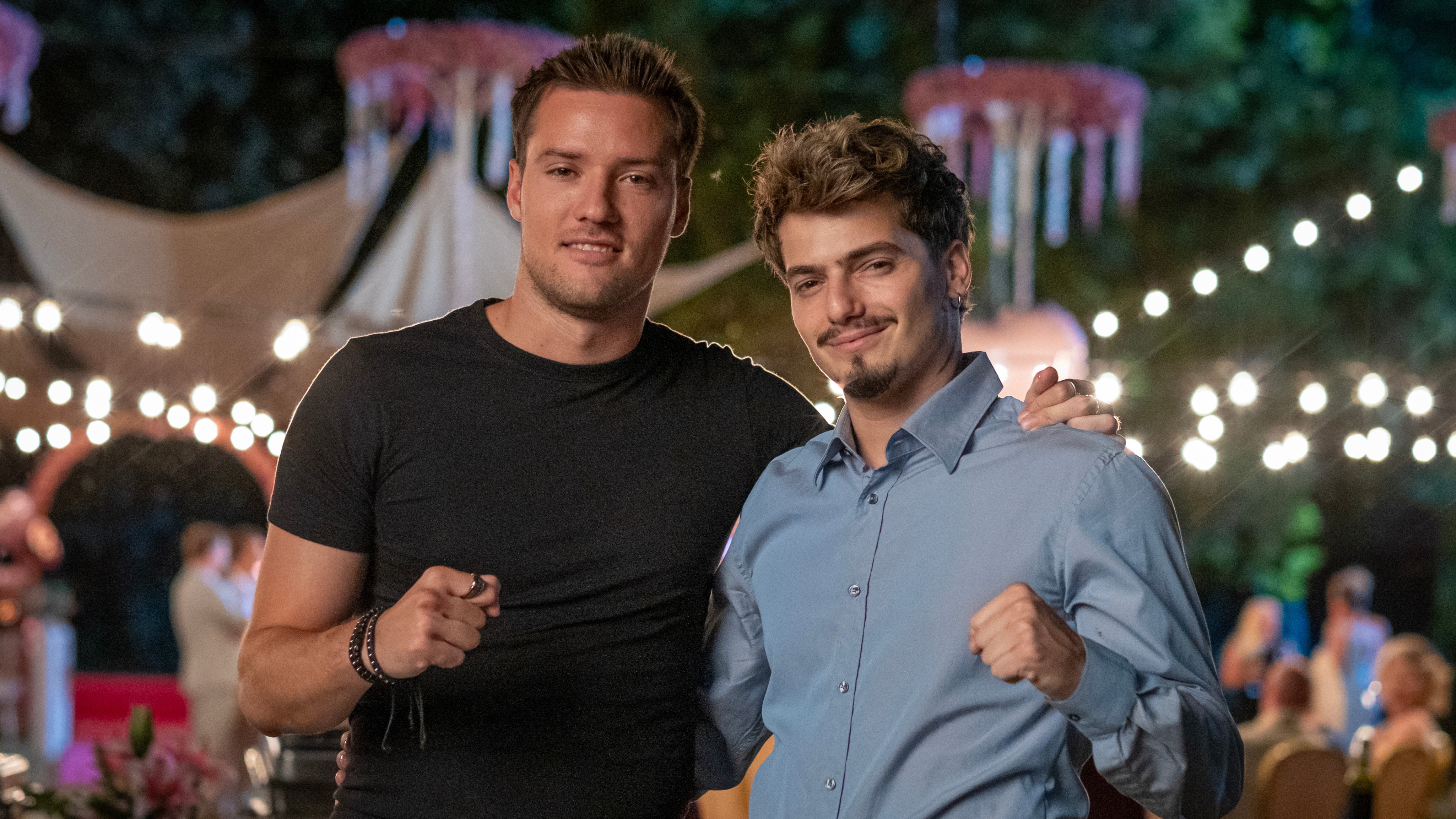 Two men posing together at night, one in a black T‑shirt and the other in a light blue shirt, smiling with arms around each other as string lights glow in the background.
