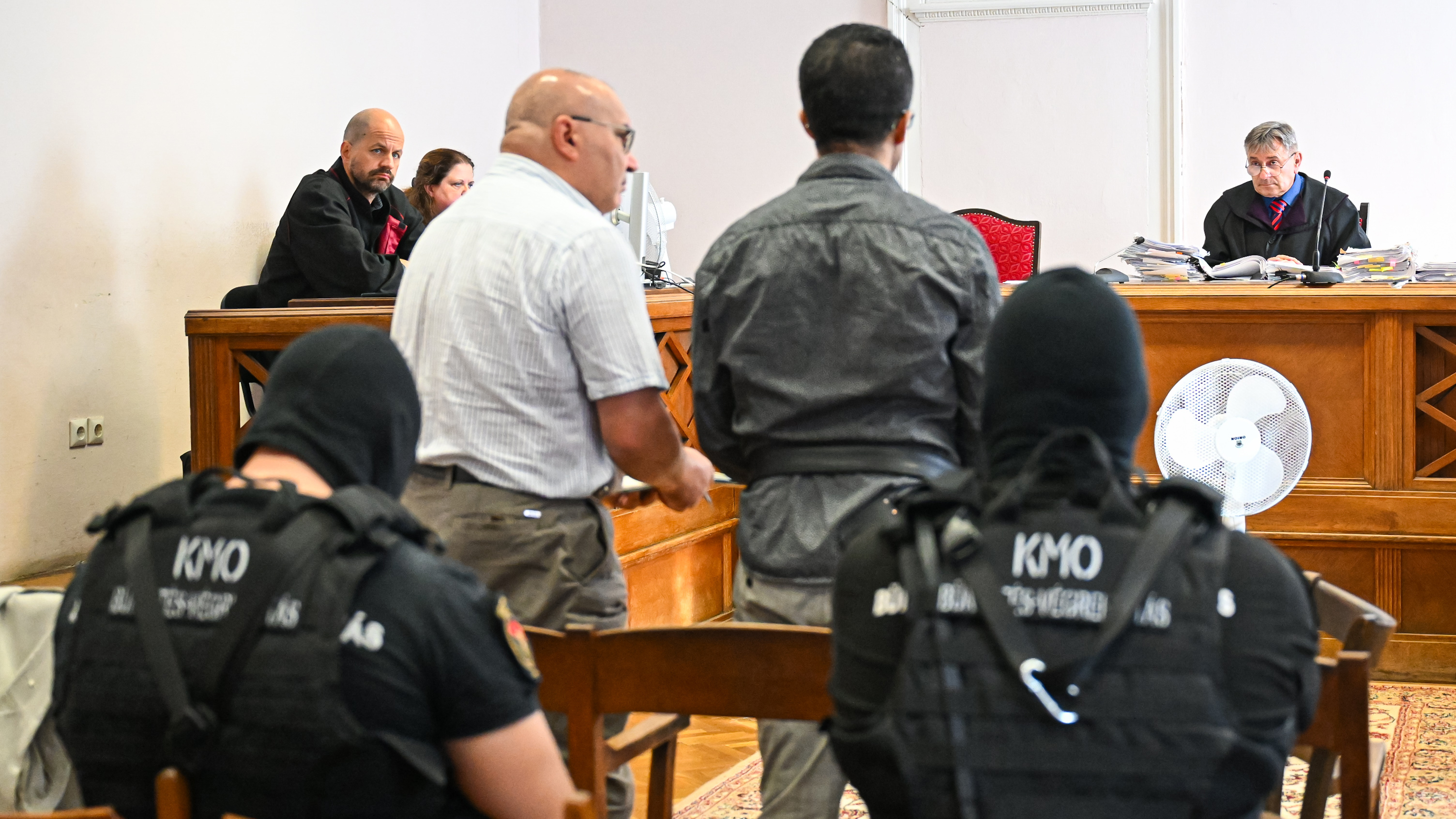 K-9 unit officers in protective vests stand in a courtroom, facing the judge at a desk cluttered with files and papers.