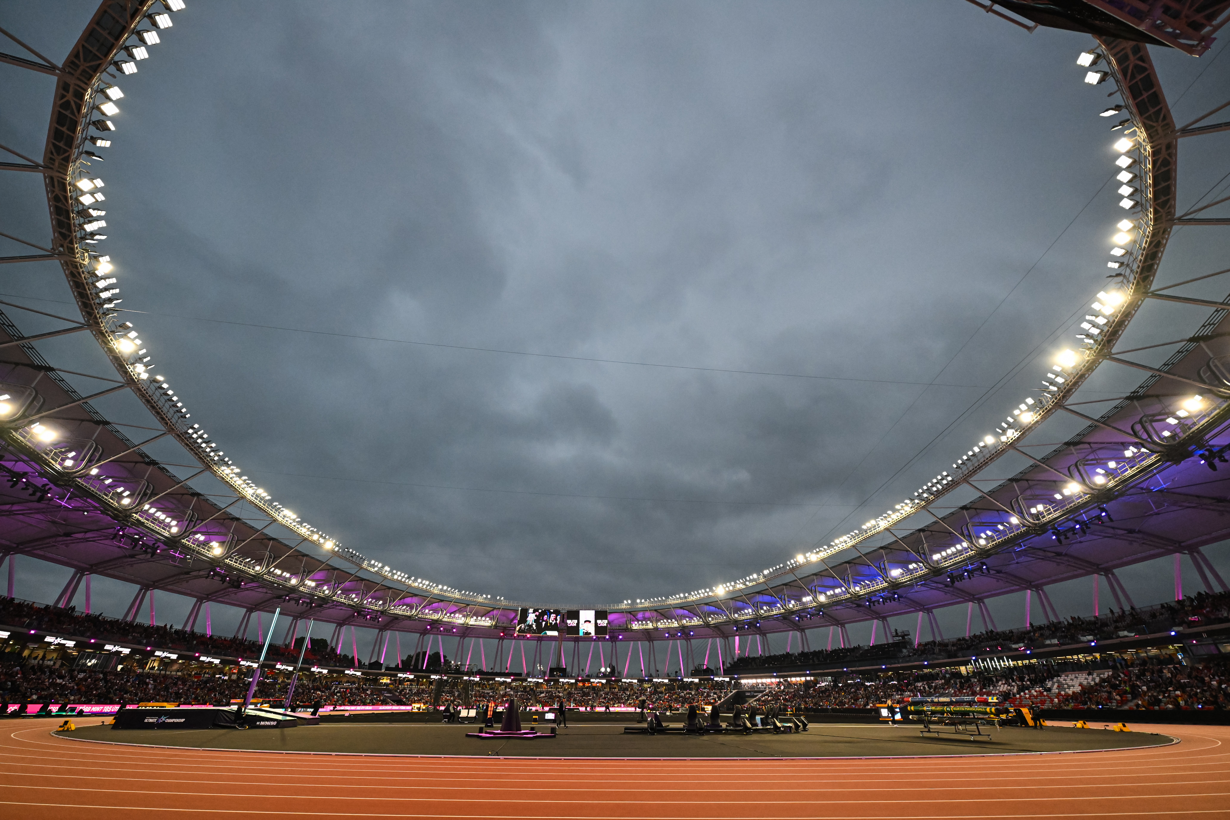 Wide view of a large outdoor stadium with a red running track, illuminated stands, and a cloudy evening sky.
