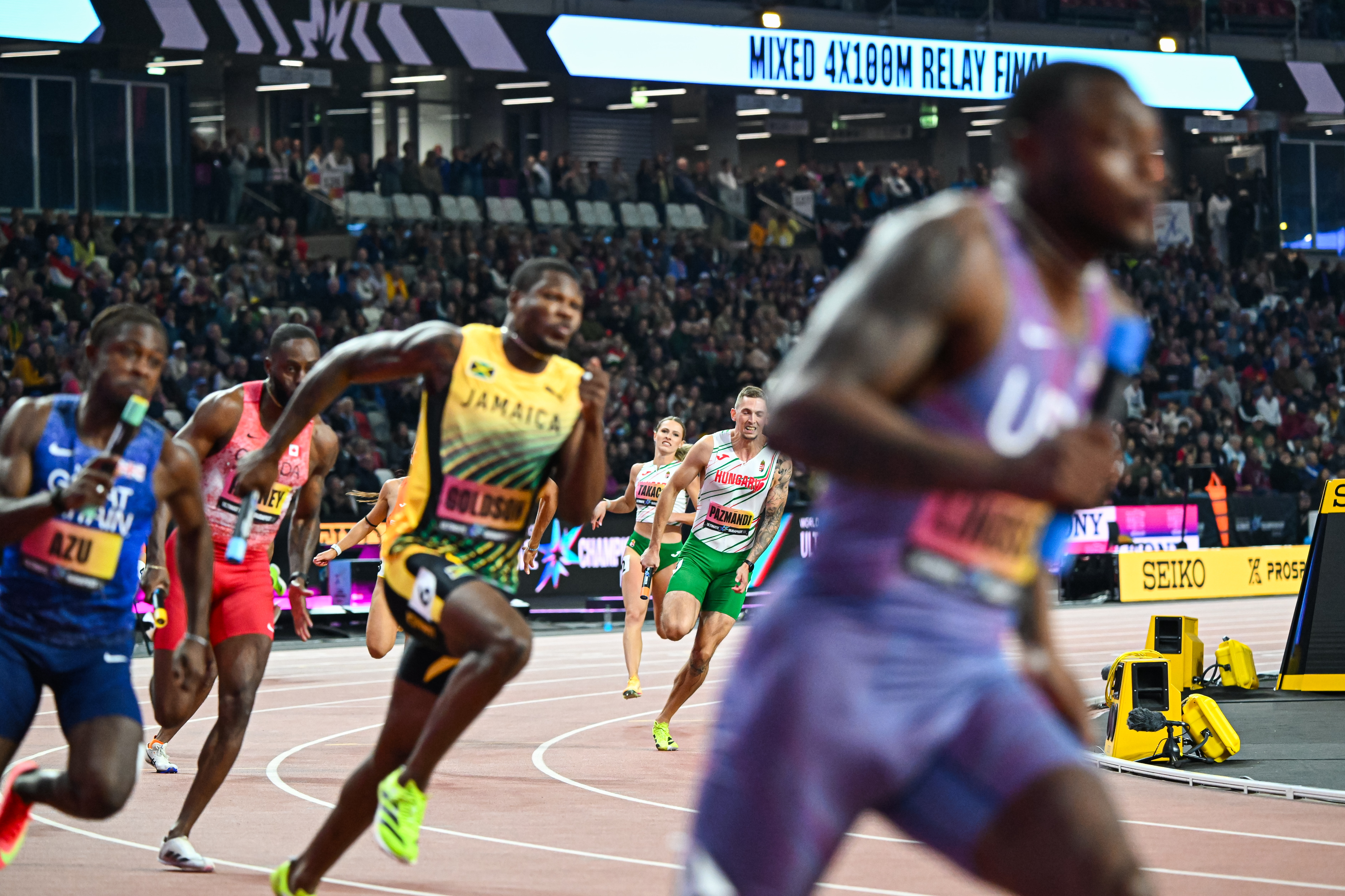 Athletes sprinting on an indoor track with a cheering crowd and banners in the stands, captured in a dynamic race scene.