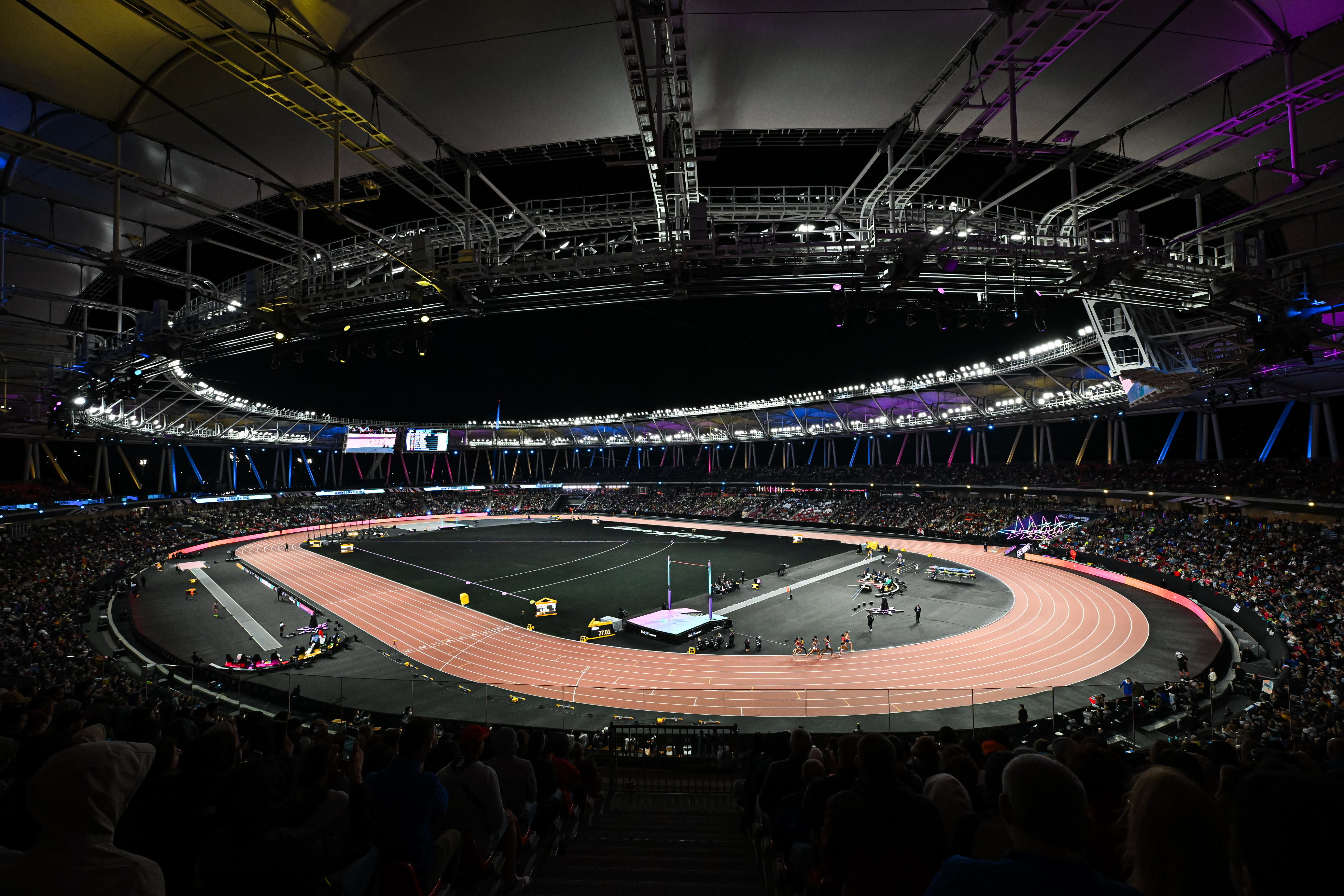 Night-time stadium scene with a circular red running track, central field, and a large audience under bright overhead rigging and lights.