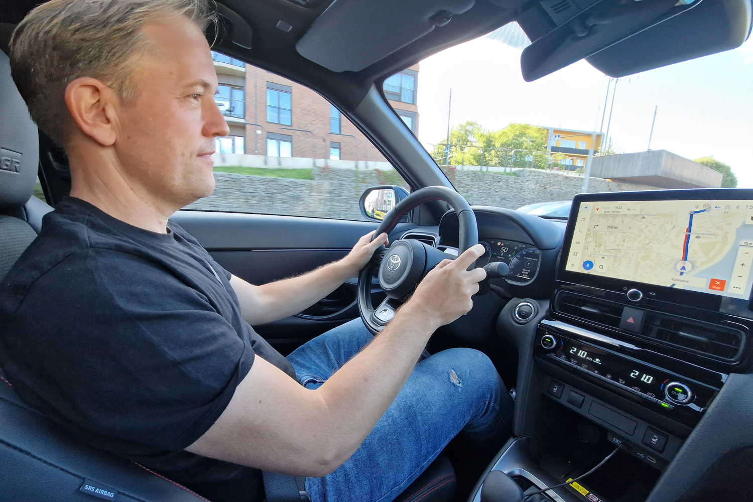 Person driving a car, hands on the steering wheel while looking ahead, with a large in-dash touchscreen showing a map on the right panel.