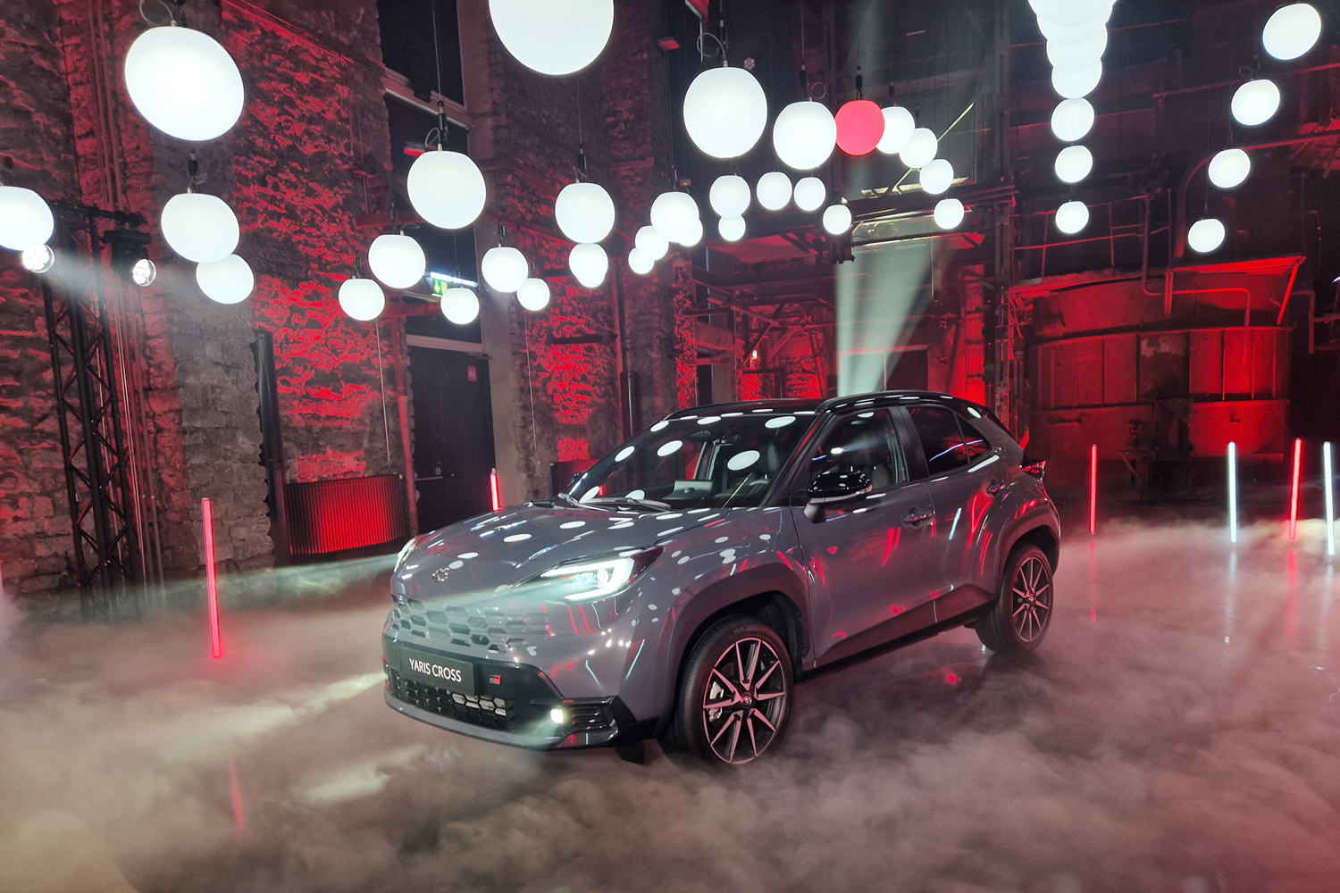 Gray Toyota Yaris Cross on a foggy showroom floor, under hanging white globe lights and red ambient lighting.