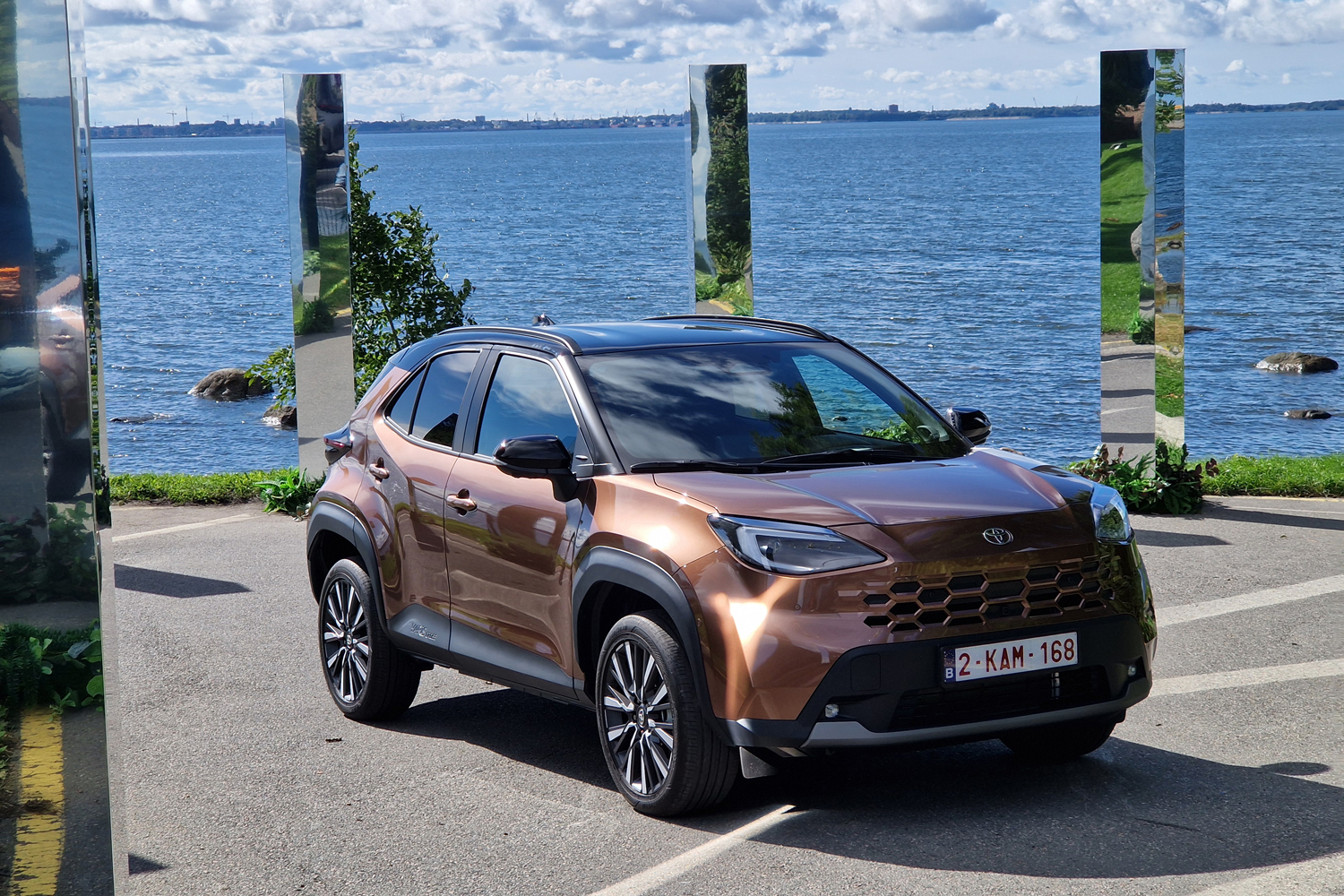 Bronze-brown Toyota SUV parked along a lakeside promenade with tall mirrored posts reflecting the scene.