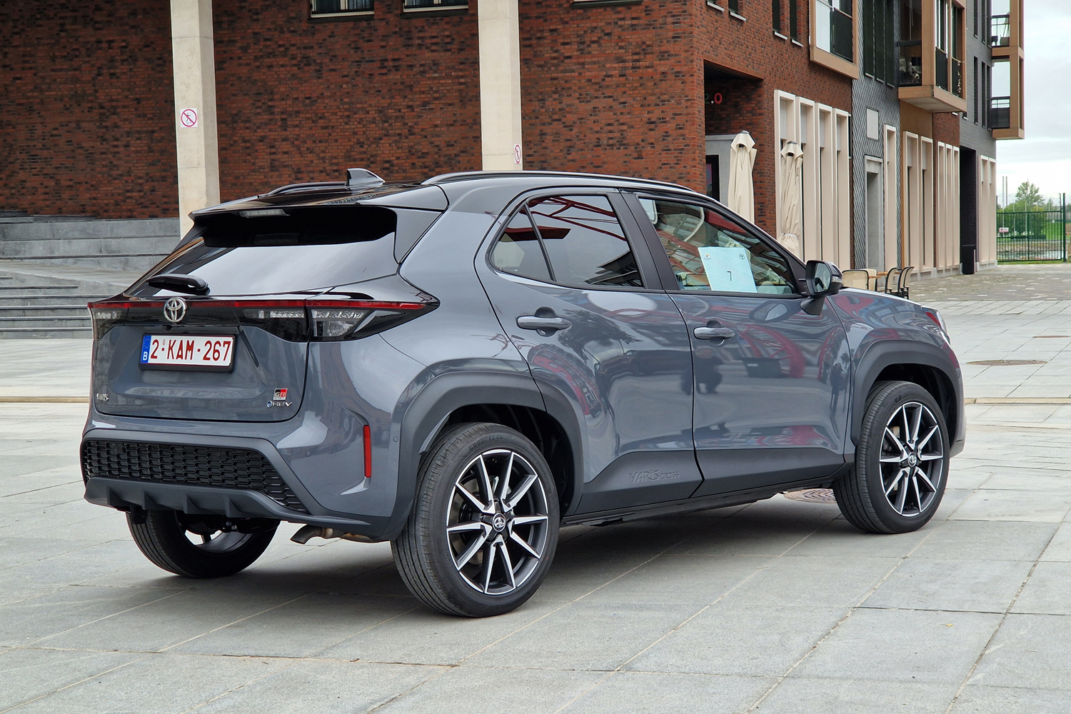 Three-quarter rear view of a gray Toyota Yaris Cross SUV parked on a paved plaza in front of a red-brick building with large windows.