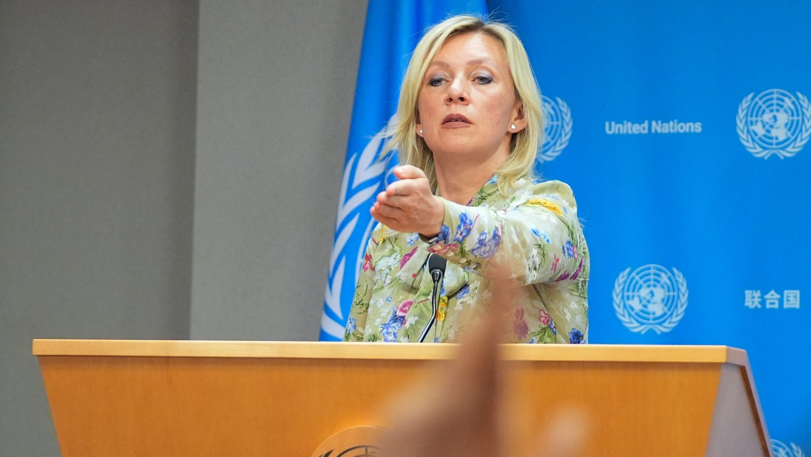 Female speaker at a podium, gesturing toward the audience with a blue United Nations backdrop behind her.
