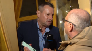 Two men face off in a doorway: a reporter with a M1 microphone asks questions to a suited man who holds a green paper in his hand.