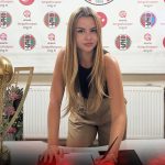 Young woman signs papers at a desk, flanked by a gold trophy on the left and a large football glove on the right, with a sports-themed backdrop.