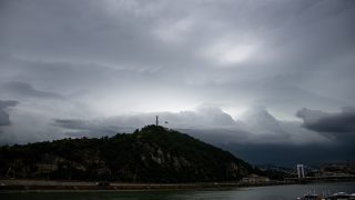 Hilltop monument with a flag on a lush hillside beneath heavy storm clouds, overlooking a river and city with a bridge in the distance.