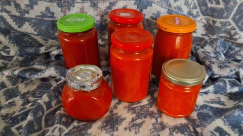 Assorted glass jars filled with red tomato sauce on a blue patterned fabric background, with colorful lids (green, red, orange, metal).